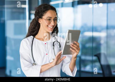 Latin American doctor woman using tablet computer, female doctor working inside office building wearing white medical coat. Stock Photo