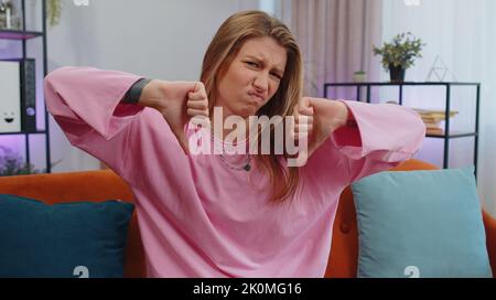Dislike. Upset girl showing thumbs down sign gesture, expressing discontent, disapproval, dissatisfied bad work at modern home apartment indoors. Displeased young woman in living room sitting on sofa Stock Photo