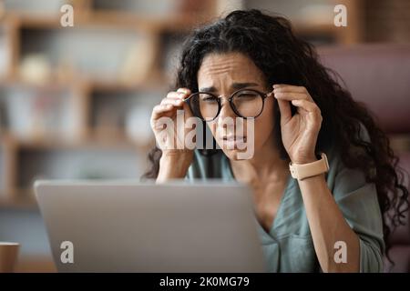 Woman sitting at desk in front of laptop, touching eyeglasses Stock Photo