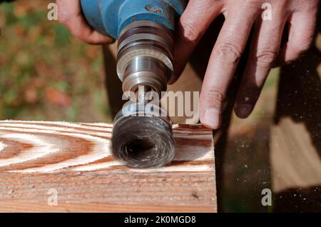 Wire brush for wood working, on a drill. Male hand is holding brushing machine electrical rotating with metal disk sanding a piece of wood. Stock Photo