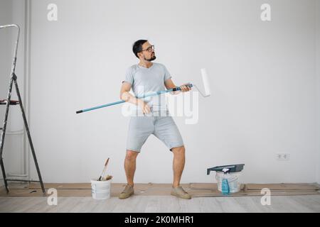 Young man, who have finally collected the necessary amount of funds for repairs, rejoice and dance. Instead of real microphone, guy using into a mop and a paint roller. Stock Photo