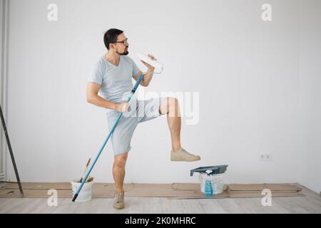 Young man, who have finally collected the necessary amount of funds for repairs, rejoice and dance. Instead of real microphone, guy using into a mop and a paint roller. Stock Photo