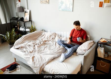 High angle view of young man sitting on bed and browsing in laptop with cup of coffee in the morning Stock Photo