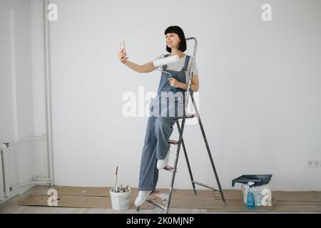 Red hair girl with cute smile holds a paint roller and smart phone in her hands. Young woman leaning on a ladder that she uses during repairs and having video call on her phone. Stock Photo