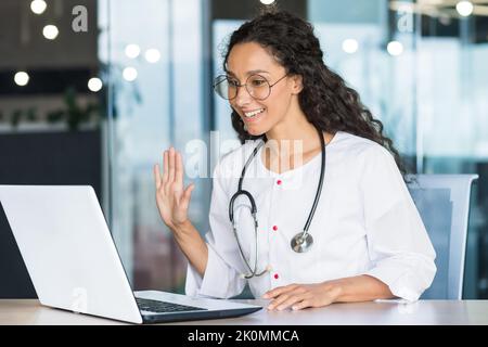 Video call online doctor consultation, latin american doctor woman with curly hair in white coat waving at laptop camera greeting patient, consulting working remotely inside clinic Stock Photo