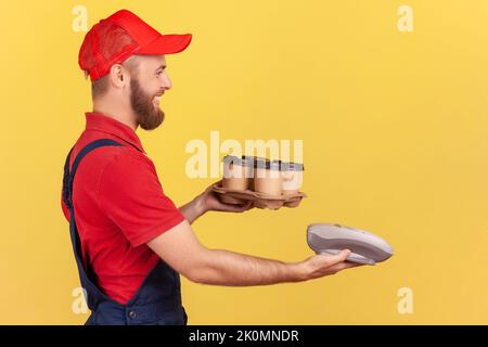 Side view portrait of delighted bearded courier man holding terminal and coffee with pizza box, bringing order, wearing overalls and cap. Indoor studio shot isolated on yellow background. Stock Photo