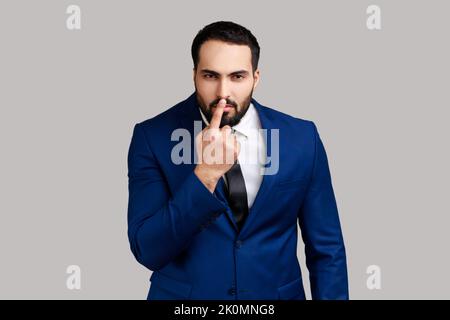 Bearded man looking with incredulous suspicious gaze and touching nose, gesturing you are liar, suspecting falsehood, wearing official style suit. Indoor studio shot isolated on gray background. Stock Photo
