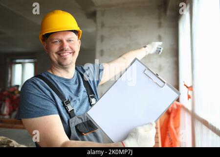 Smiling builder worker in hard hat holding blank sheet of paper on clipboard Stock Photo