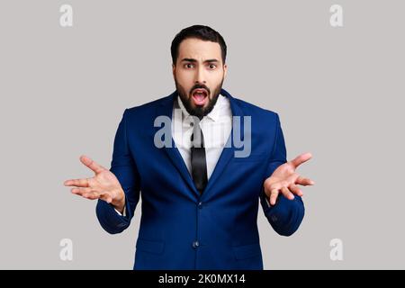 Portrait of bearded man standing with raised hands and indignant expression, asking reason of conflict, wearing official style suit. Indoor studio shot isolated on gray background. Stock Photo