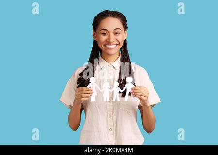 Portrait of happy woman with black dreadlocks holding paper chain people in hands, concept of happy family, parenthood, childhood, wearing white shirt. Indoor studio shot isolated on blue background. Stock Photo