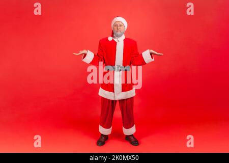 Full length portrait of elderly man with gray beard wearing santa claus costume shrugging shoulders as doesn't know answer, being uncertain, not sure. Indoor studio shot isolated on red background. Stock Photo