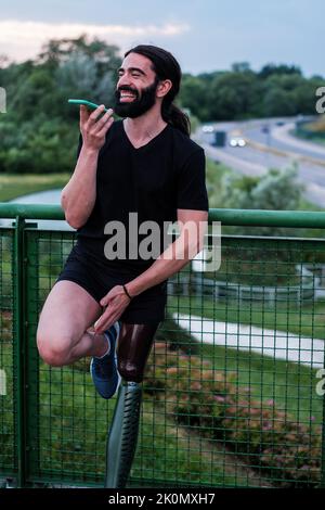 Young man with long beard hair talking on the smartphone outdoors. Prosthesis Stock Photo