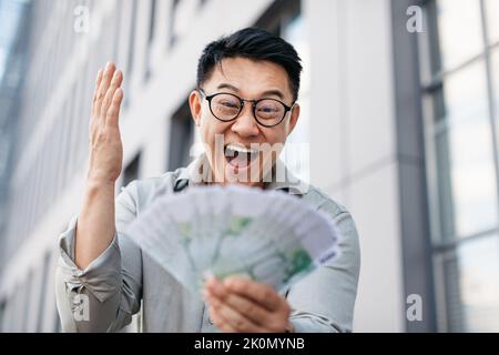 Big win concept. Overjoyed asian man holding fan of American money, celebrating increase in profit, standing outdoors Stock Photo