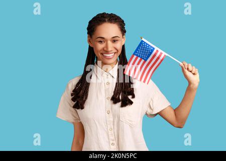 Smiling patriotic woman with dreadlocks holding in hand flag of united states of america, celebrating independence day, wearing white shirt. Indoor studio shot isolated on blue background. Stock Photo