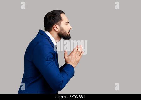 Side view of man holding hands in prayer, looking with imploring pleading expression, begging help, asking forgiveness, wearing official style suit. Indoor studio shot isolated on gray background. Stock Photo