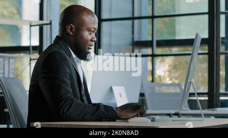Side view african american senior businessman analyst ceo manager sit at office desk typing email chatting on corporate computer concentrated search Stock Photo