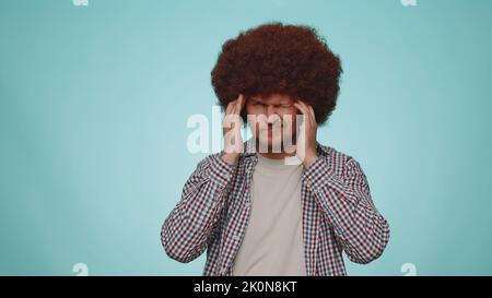 Bearded young adult man in shirt rubbing temples to cure headache problem, suffering from tension and migaine, stress, grimacing in pain, high blood pressure isolated alone on blue wall background Stock Photo