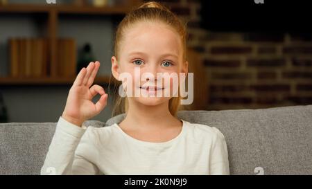 Portrait of caucasian happy child girl smiling kid sitting on sofa at room looking at camera showing ok gesture everything fine like sign positive Stock Photo