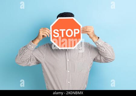 Portrait of man covering face with Stop symbol, anonymous person holding red traffic sign, warning to go, prohibition concept, wearing striped shirt. Indoor studio shot isolated on blue background. Stock Photo