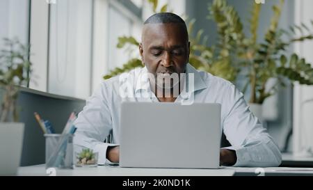 Concentrated african man employee american expert focused specialist sits in office works on laptop use personal computer typing email chatting Stock Photo