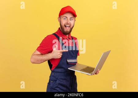 Side view of excited worker man wearing blue overalls, red T-shirt and cap working on laptop, pointing at screen with finger, looking at camera. Indoor studio shot isolated on yellow background. Stock Photo