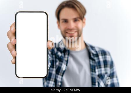 Defocused photo of a caucasian young man, looks at camera, hold in hand smartphone with white blank mock-up screen for presentation or advertising, stand on isolated white background, smiles Stock Photo