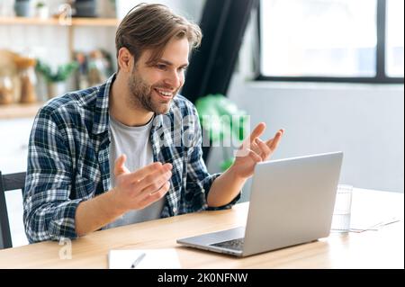 Video communication. Friendly caucasian young man, company employee, freelancer, sit at home in kitchen, works remotely uses laptop, talking on video conference with client or colleague, smiles Stock Photo
