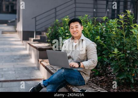 Happy mature asian man working on laptop computer outdoors, sitting on bench in urban area and using pc Stock Photo