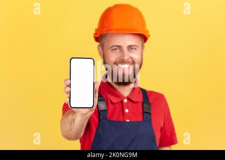 Portrait of happy smiling builder man wearing blue uniform and protective helmet holding smart phone with blank screen for advertisement. Indoor studio shot isolated on yellow background. Stock Photo