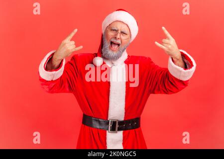 Excited elderly man with gray beard wearing santa claus costume shouting and showing rock and roll sign, looking at camera, celebrating Christmas. Indoor studio shot isolated on red background. Stock Photo