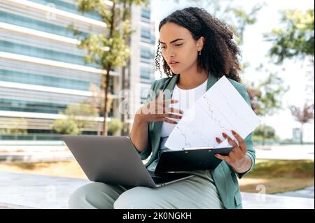 Busy focused latino or brazilian business woman, financial analyst, entrepreneur, trader, sitting outdoors with laptop, focusing on analyzing financial documents, checking reports and graphs Stock Photo