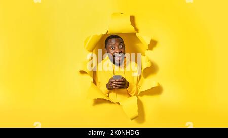 Overjoyed black man looking with open mouth, using cellphone and surfing internet, torn hole in yellow background Stock Photo