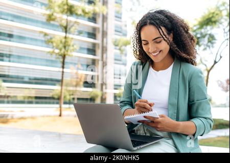Online webinar. Smart successful brazilian or latino young woman, university student, freelancer, sit outdoors with laptop, listening to an online lecture, writing down information in notebook, smiles Stock Photo