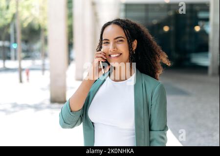 Wireless communication. Busy positive latino or brazilian woman, successful realtor, business lady in elegant clothes, has a pleasant conversation on a cell phone while standing outdoors, smiling
