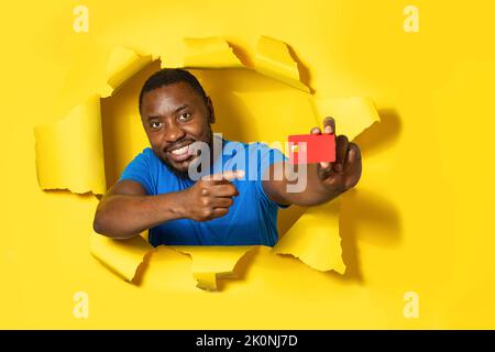 Happy african american man showing credit bank card and pointing on it isolated through torn yellow paper background Stock Photo