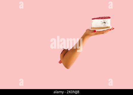 Creative Halloween minimal concept with cheesecake, spider and woman's hand. Pastel pink torn paper. Creative spooky holiday food concept. Copy space. Stock Photo