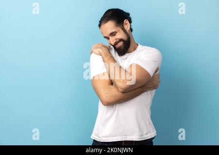 Portrait of happy complacency handsome man with beard wearing white T-shirt standing and hugging himself with closed eyes and toothy smile enjoying. Indoor studio shot isolated on blue background. Stock Photo