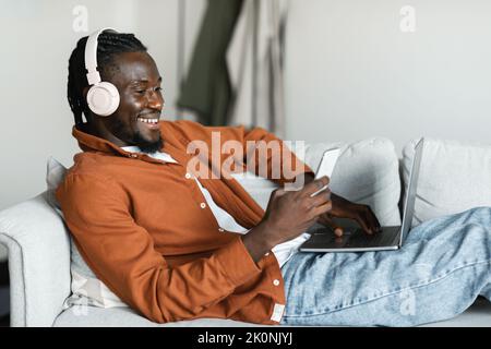 Smiling black man lying on couch with pc, using cellphone and wearing headset, taking break from remote work Stock Photo
