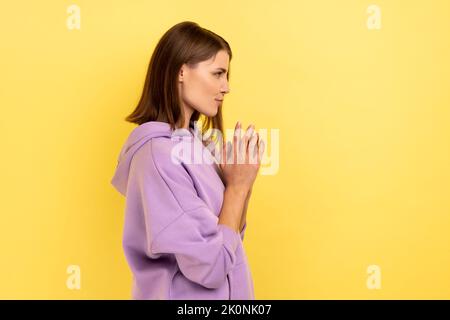 Side view of devious cunning young woman clasping hands and smirking mysteriously, scheming cheats, evil prank, wearing purple hoodie. Indoor studio shot isolated on yellow background. Stock Photo