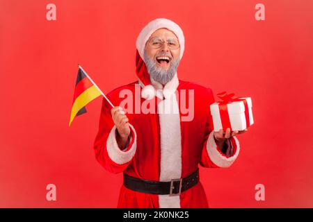 Portrait of happy excited elderly man with gray beard wearing santa claus costume holding German flag and box with Christmas present. Indoor studio shot isolated on red background. Stock Photo