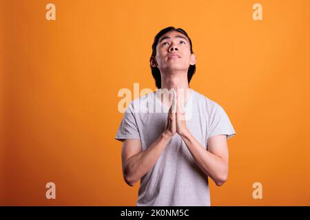 Young man praying folding hands, faith, worship concept. Religious asian teenager pleading, looking upwards with hope, person praising gods, medium shot on orange background Stock Photo