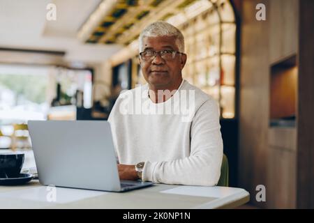 Senior businessman looking at the camera while working in a modern cafe. Mature businessman using a laptop while working remotely. Stock Photo