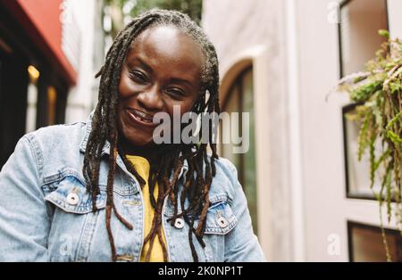 Ethnic woman with dreadlocks smiling happily while standing outdoors. Cheerful senior woman going out in casual clothing. Stock Photo