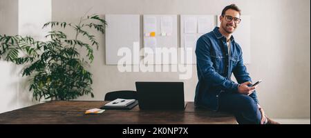 Successful businessman smiling at the camera while sitting on a desk with a smartphone. Happy businessman with eyeglasses working remotely in a creati Stock Photo