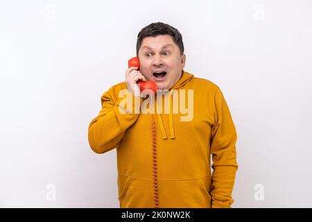 Portrait of happy man talking landline telephone holding in hand handset, keeping mouth open, hearing astonishing news, wearing urban style hoodie. Indoor studio shot isolated on white background. Stock Photo