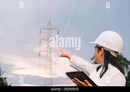 Asian female electrical engineer working on laptop near high voltage pole, inspecting power grid. Stock Photo
