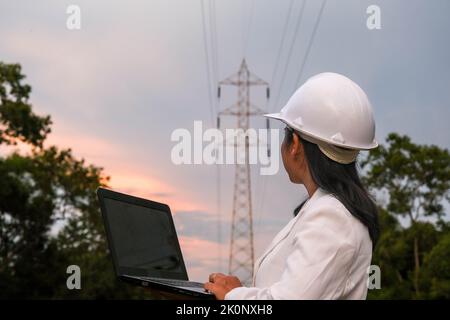 Asian female electrical engineer working on laptop near high voltage pole, inspecting power grid. Wind Turbine Engineer for Electric Power Generation. Stock Photo