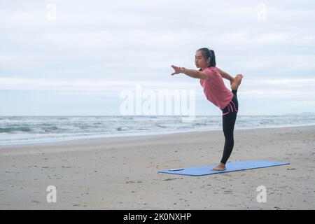 Asian woman practicing yoga at seashore. Young beautiful woman stretching and doing yoga outdoors on blue yoga mat on beach. relaxing in nature Stock Photo