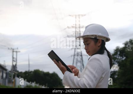 Asian female electrical engineer working on tablet near high voltage pole, inspecting power grid. Wind Turbine Engineer for Electric Power Generation. Stock Photo