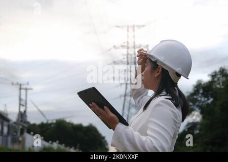 Asian female electrical engineer working on laptop near high voltage pole, inspecting power grid. Wind Turbine Engineer for Electric Power Generation. Stock Photo